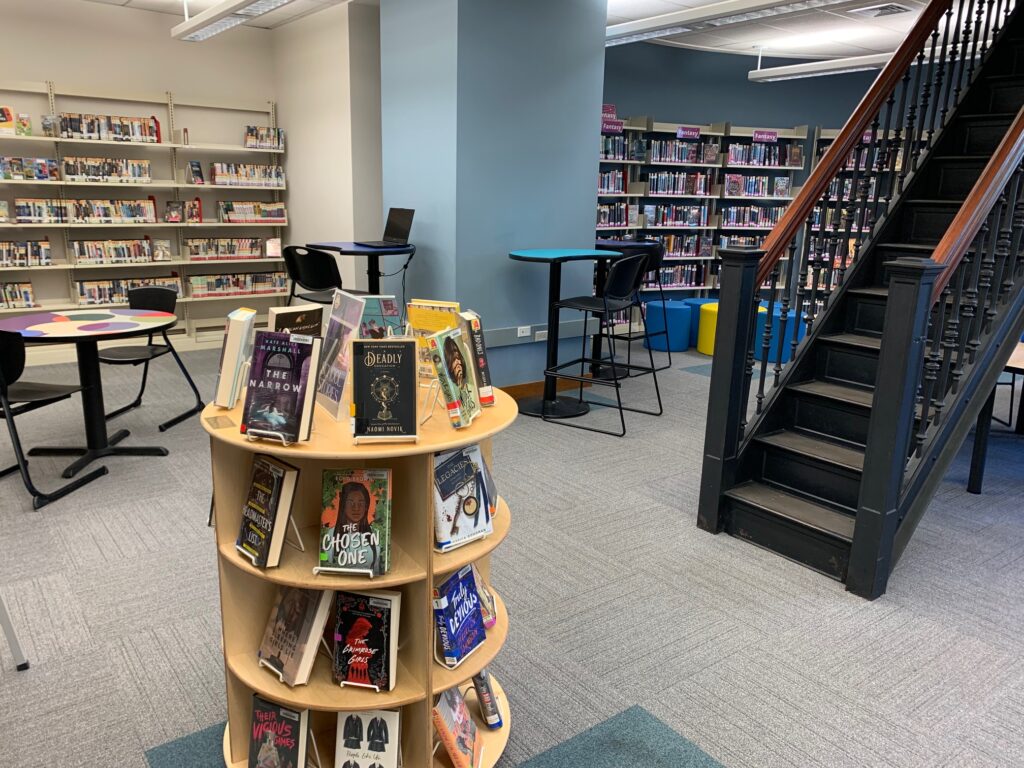 A picture of the teen room. There are tables, a book display, and a staircase.
